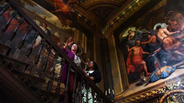 Two visitors standing on an ornate dark wood staircase surrounded by large colourful classical paintings covering the walls